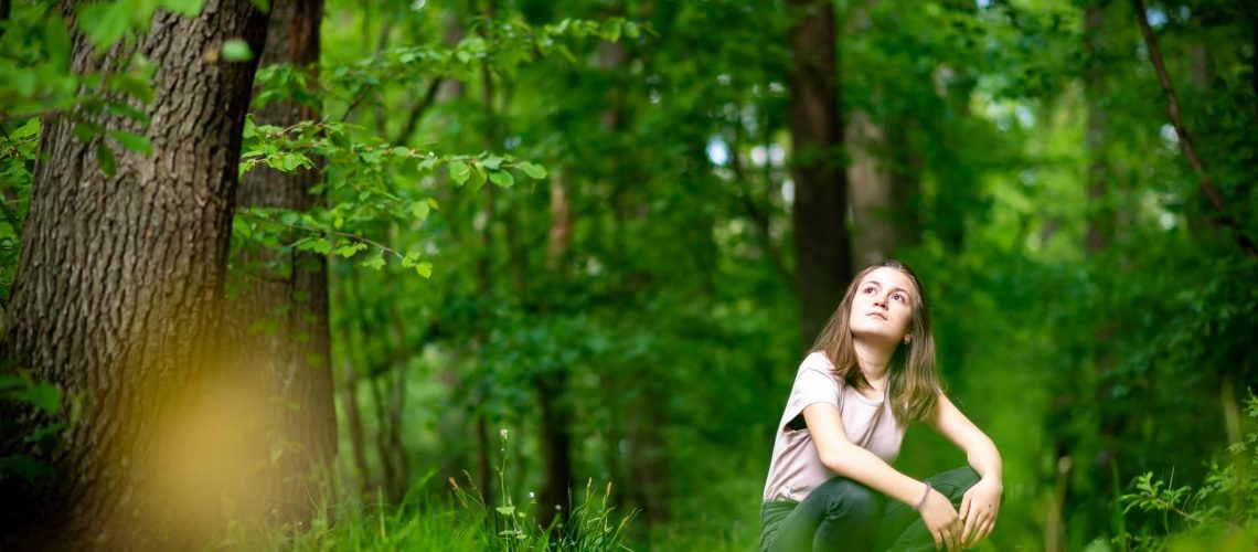 Junge Frau sitzt im Wald und sieht nachdenklich nach oben